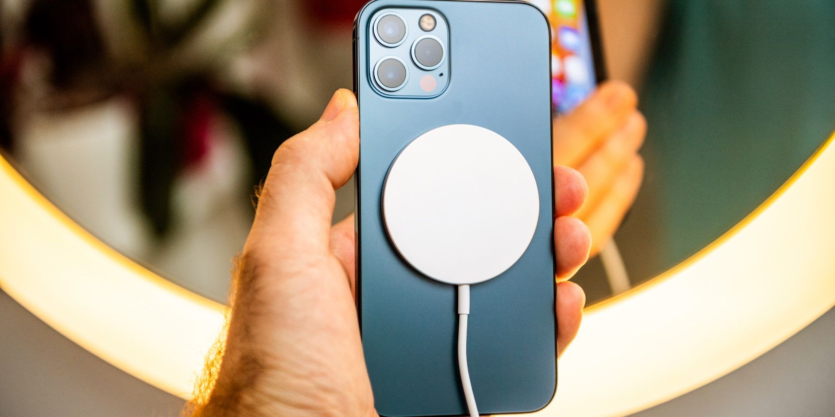 دست مردی که جلوی آینه می گیرد مدل جدید گوشی هوشمند آیفون 12 پرو مکس 5G توسط Apple Computers از نمای نزدیک دستگاه تلفن همراه آبی اقیانوس آرام در حال شارژ با MagSafe