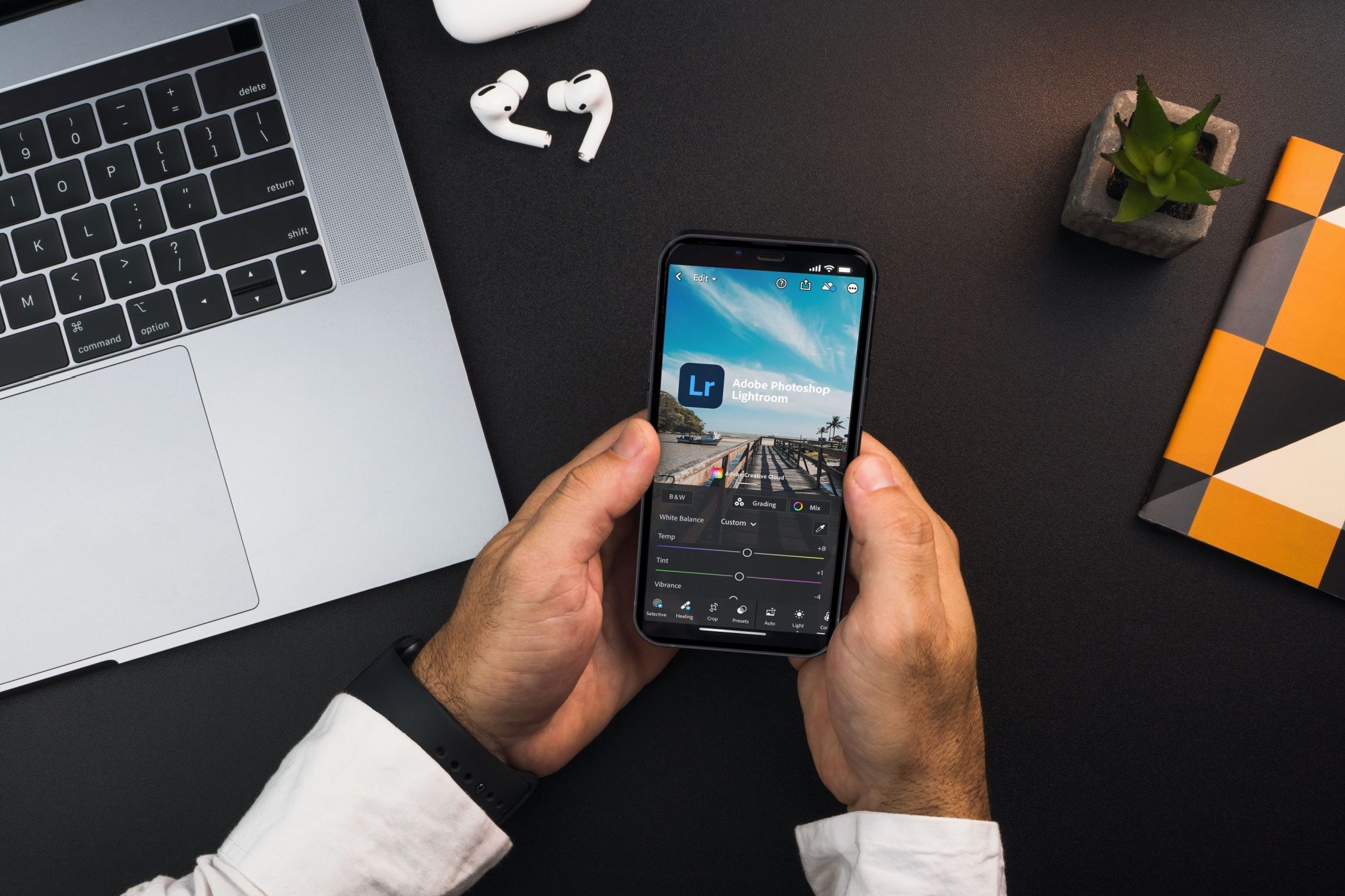 Man Holding Smartphone Adobe Photo ویرایش عکس Adobe ، با لپ تاپ ، گوشواره و نوت بوک در پس زمینه.