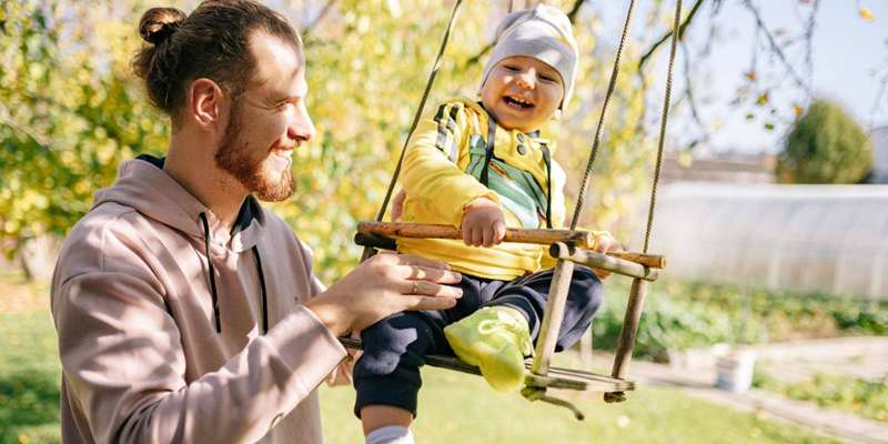 مردی در کتم خاکستری که پسری با تی‌شرت سبز را در تاب صندلی نگه داشته است