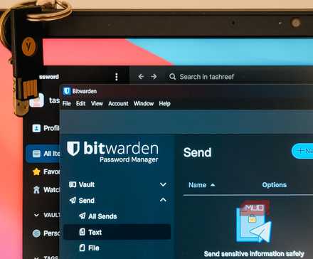 برنامههای Bitwarden و 1Password در پنجرههای همپوشان بر روی ویندوز ۱۱ باز هستند و یک YubiKey در بالای درپوش لپتاپ آویزان است