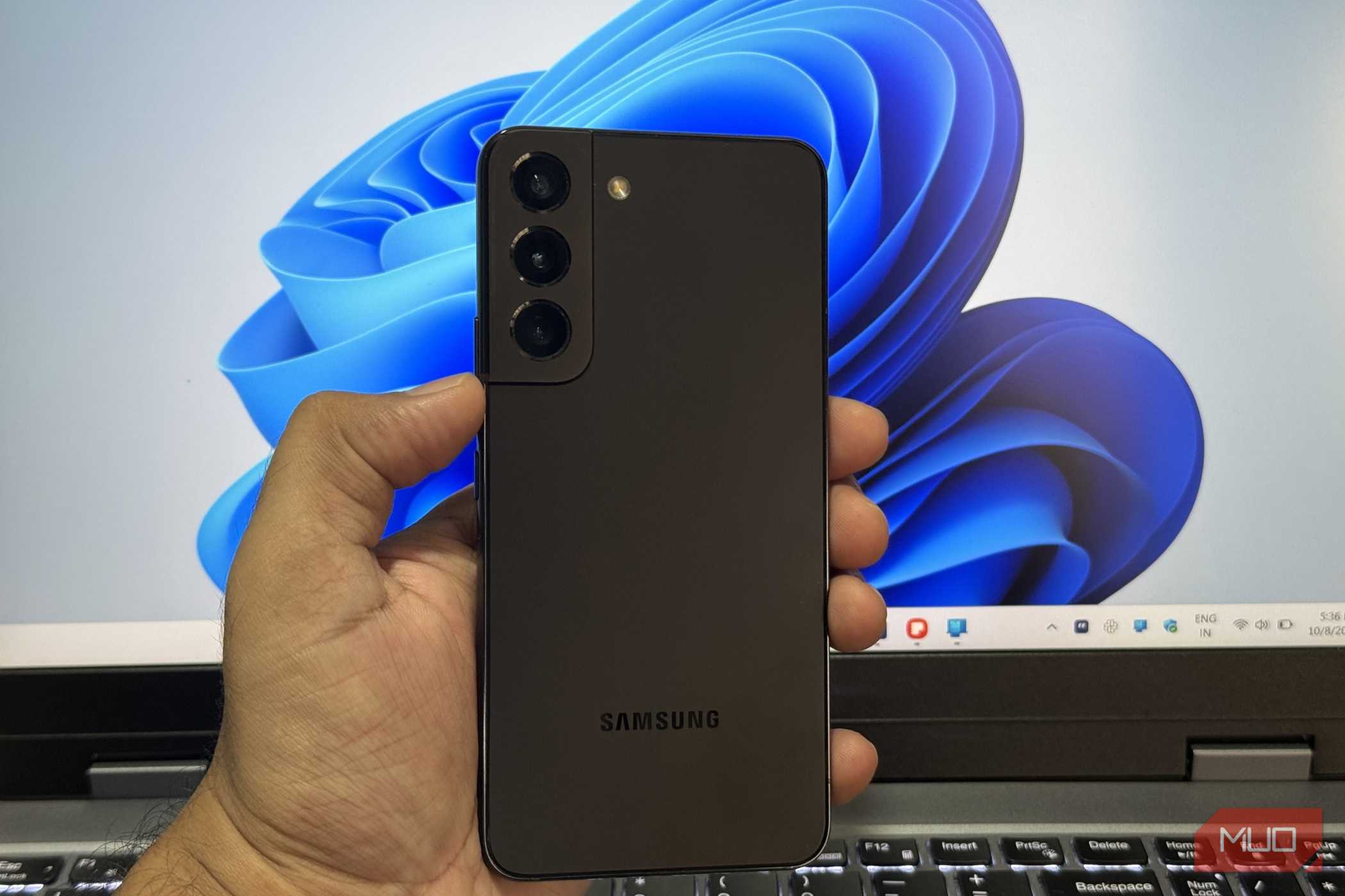 گوشی سامسونگ S22 در دست با لپ‌تاپ ویندوز در پس‌زمینه